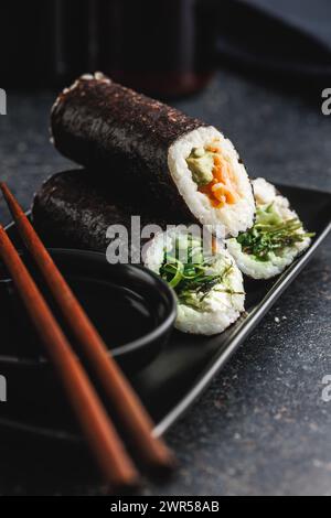 Une assiette noire avec des morceaux de sushi avec des baguettes et un bol de sauce soja. Le sushi montre une variété de couleurs, de textures et de garnitures, showc Banque D'Images