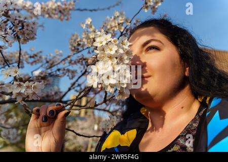 Attrayant plus de taille jeune femme reniflant branche de fleur de cerisier dans le parc au printemps Banque D'Images