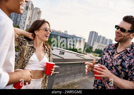 amis joyeux et interraciaux dans des attitudes vibrantes buvant dans des tasses rouges à la fête sur le toit ensemble Banque D'Images