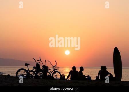 Quatre surfeurs et leurs vélos et planches sont silhouettés par le soleil couchant sur la plage sur la rive nord d'Oahu. Banque D'Images