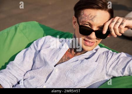 joyeux homme beau dans des vêtements décontractés avec des tatouages et des lunettes de soleil élégantes souriant à la caméra sur le toit Banque D'Images