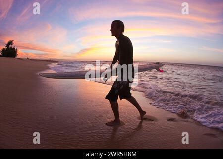 Un jeune homme quittant le surf, sous un beau coucher de soleil sur la rive nord. Oahu, Hawaï. Banque D'Images