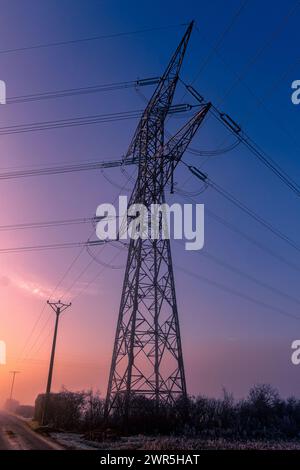 Pylône métallique transportant de l'électricité haute tension avec un ciel bleu et rouge, couleurs de l'aube. France, Europe Banque D'Images