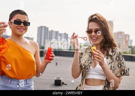 femmes joyeuses attrayantes dans des tenues décontractées vibrantes avec des lunettes de soleil soufflant des bulles de savon sur le toit Banque D'Images