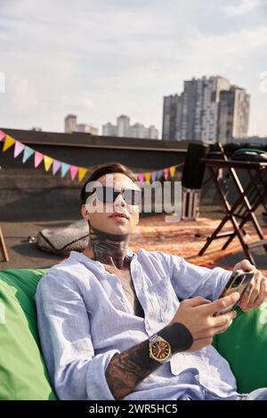 joyeux homme avec des tatouages et des lunettes de soleil se relaxant avec le téléphone à la main et regardant la caméra Banque D'Images