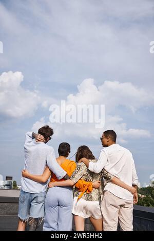 vue arrière d'un groupe d'amis interraciaux dans des vêtements vibrants confortables posant sur le toit ensemble Banque D'Images