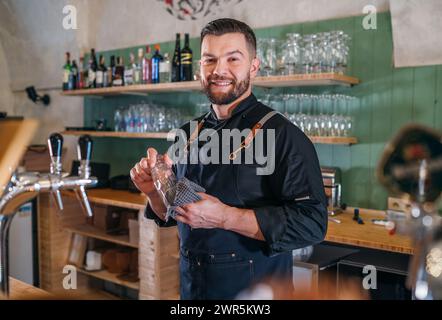 Portrait d'heureux barman barbu souriant vêtu d'un uniforme noir avec un tablier essuyant le verre à bière au comptoir du bar. Des gens qui réussissent, un travail acharné, Banque D'Images