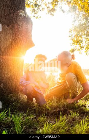 Cette image évocatrice capture une interaction réconfortante entre un grand-père et son petit-enfant dans la douce lumière dorée du soleil couchant. L'homme plus âgé, avec une barbe blanche et portant une chemise jaune, accroupit pour être à hauteur des yeux avec le jeune enfant, qui est également accroupi. Ils sont près d'un grand arbre, et le soleil s'illumine à travers les branches, les entourant d'une atmosphère chaude et lumineuse. Le grand-père semble partager la sagesse ou une histoire avec l'enfant, qui écoute attentivement. C'est une image du lien générationnel et des moments simples et précieux qui forment le tissu de la famille moi Banque D'Images