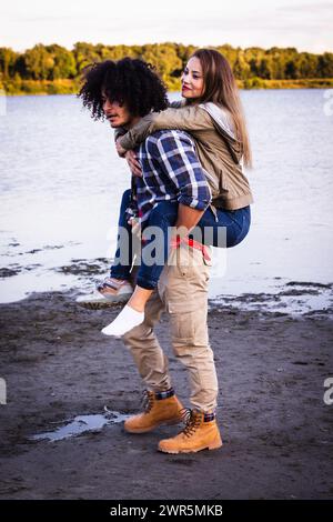 Capturée dans un cadre de canevas au bord d'un lac, cette image dynamique montre un couple multiracial engagé dans une balade ludique en piggyback. Un homme afro-américain avec les cheveux noirs bouclés et une chemise à carreaux porte une femme caucasienne avec de longs cheveux blonds et une veste beige. La femme, portant un Jean denim, a les bras enroulés autour des épaules de l'homme, affichant un large sourire, révélateur de plaisir et d'affection. L'homme, vêtu d'un pantalon kaki et de bottes brunes, montre une expression concentrée, indiquant peut-être l'effort de porter son compagnon. Le fond révèle un lac serein et des arbres lointains, contri Banque D'Images