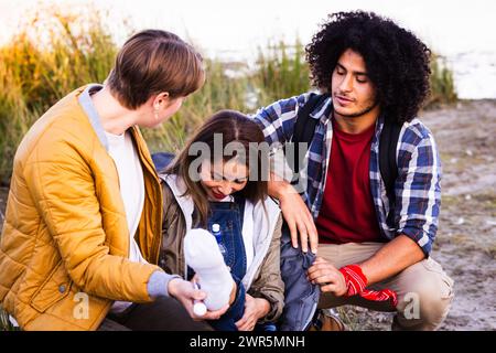 Dans cette image, un moment de soin se déroule dans la tranquillité de la nature comme un groupe d'amis assiste à une partie blessée. Une femme, apparemment dans la douleur, est le sujet de leur préoccupation. Un ami, une personne aux cheveux courts dans une veste jaune, étend une bouteille d'eau à la femme, indiquant un geste d'aide et de confort. Ils sont accompagnés par un homme aux cheveux bouclés et à la barbe, portant une chemise à carreaux, qui semble empathique et prêt à aider. La composition diversifiée du groupe, comprenant une femme caucasienne et un homme d'ascendance moyen-orientale ou nord-africaine, reflète une dynamique multiethnique. Le plein air Banque D'Images