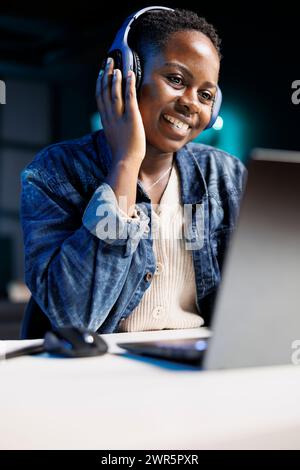 Femme noire joyeusement en utilisant un ordinateur portable et un casque sans fil. Influenceuse afro-américaine multitâche, assistant à une réunion en ligne tout en écoutant de la musique dans son casque. Banque D'Images