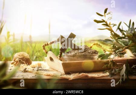 Croix chrétienne sur cendres dans un récipient en verre sur un plateau en bois sur une table table avec des feuilles d'olivier dans la nature. Vue de face. Banque D'Images