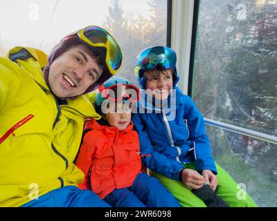 Des sourires brillent sur les visages de trois skieurs, un homme adulte et de jeunes garçons, alors qu'ils prennent une photo sur un skilift, avec des arbres givrés, la piste de ski sous t Banque D'Images