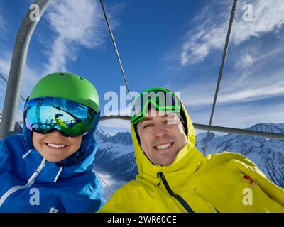 Une paire de skieurs heureux adulte et un enfant, portant des vestes de ski lumineuses, des lunettes, capturent un moment joyeux sur un skilift avec les pics blancs derrière Banque D'Images