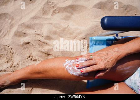 Une femme applique de la crème solaire sur ses jambes tout en prenant un bain de soleil dans une chaise de plage. Prévention du cancer de la peau. Positivité corporelle Banque D'Images