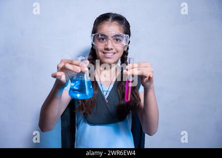 Heureuse jeune fille indienne en uniforme scolaire portant des lunettes de sécurité de laboratoire tout en travaillant sur son projet de chimie expérience avec des tubes à essai et des fla coniques Banque D'Images