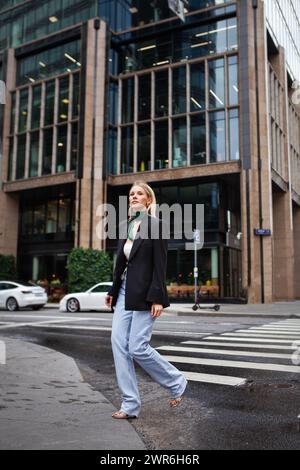 Belle et élégante jeune femme traversant la route à piéton dans la ville habillée à la mode en jeans et blazer. Style de rue de mode sur fond de b Banque D'Images