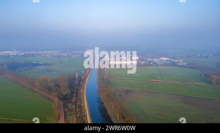 Une scène matinale brumeuse se déroule, révélant la juxtaposition des champs agricoles et du développement industriel par le haut. Vue surélevée du canal brumeux divisant les terres agricoles rurales et la zone industrielle. Photo de haute qualité Banque D'Images