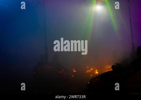 Cette image représente l'atmosphère animée et brumeuse d'un champ de foire, en se concentrant sur les voitures pare-chocs au milieu d'un dense effet de brouillard. Les faisceaux lumineux vibrants de la structure suspendue coupent la brume, réfléchissent les surfaces et créent une lueur éthérée. Les formes obscurcies des personnes et des voitures renforcent la sensation dynamique et festive d'une nuit à la foire. Voitures pare-chocs Fairground dans une ambiance brumeuse. Photo de haute qualité Banque D'Images