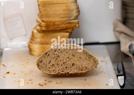 L'image capture une demi-tranche de pain à grains entiers reposant sur une planche à découper en bois avec des miettes autour, suggérant que le processus de tranchage vient de se produire. En arrière-plan, les tranches empilées de pain blanc sont légèrement floues, contrastant avec le morceau de grain entier au premier plan. Un couteau se trouve sur le côté, indiquant que le pain a été fraîchement coupé, probablement dans une cuisine de maison ou de boulangerie. Pain de grains entiers tranché sur une planche à découper. Photo de haute qualité Banque D'Images