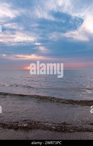 Cette photographie capture la majesté tranquille d’un coucher de soleil sur la mer, avec le soleil plongeant sous l’horizon, sa lumière diffusée par un voile de nuages. Le ciel est une toile de bleus doux et de roses subtils, reflétant le salut final du soleil à la journée. La mer, douce et tamisée, chevauche le rivage rocheux, contribuant à l'atmosphère sereine. Les nuages sont artistiquement striés à travers le ciel, ajoutant de la profondeur et de la texture à l'affichage céleste. Cette scène évoque un sentiment de calme et d’introspection alors que le jour passe à la nuit. Le délice du crépuscule : coucher de soleil tranquille sur la mer douce. Photo de haute qualité Banque D'Images