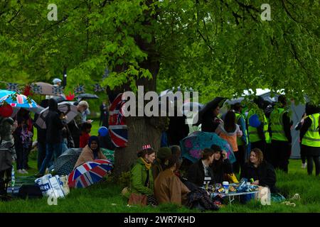 Les gens s'abritent de la pluie sous un arbre alors qu'ils se rassemblent pour voir le couronnement du roi Charles III à l'écran à Hyde Park, Londres. Banque D'Images