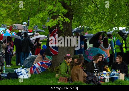 Les gens s'abritent de la pluie sous un arbre alors qu'ils se rassemblent pour voir le couronnement du roi Charles III à l'écran à Hyde Park, Londres. Banque D'Images