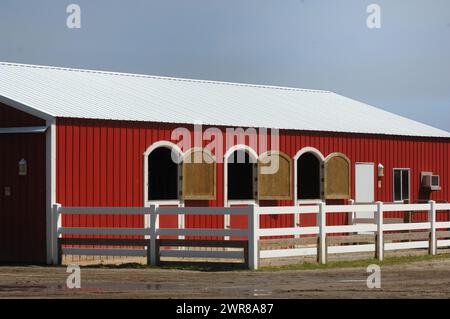 Grange rouge côté aluminium avec fenêtres incurvées et portes ouvrant sur l'extérieur grange joliment entretenue sur la ferme rouge et blanc avec clôture en PVC blanche sur la petite campagne Banque D'Images