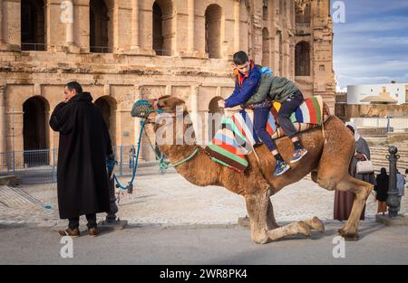 Deux jeunes garçons font une promenade à dos de chameau à l'extérieur de l'amphithéâtre romain El Jem (Thysdrus), bien conservé, en Tunisie. Banque D'Images