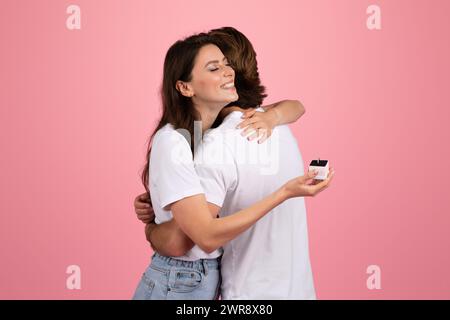 Femme rayonnante embrassant un homme qui tient une bague de fiançailles dans une boîte derrière son dos Banque D'Images