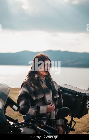 Une femme Latina contemplative se tient à côté de sa moto, regardant au loin par un lac serein avec des montagnes en toile de fond Banque D'Images