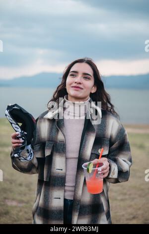 Une jeune femme souriante tient des lunettes de motocross et un cocktail frais, debout dans un champ ouvert avec un lac en arrière-plan Banque D'Images