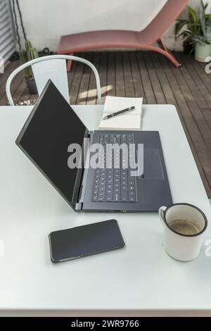 Une petite table de terrasse en métal peinte en bleu Uni, avec un ordinateur portable ouvert sur le dessus avec une tasse de café, un smartphone et un bloc-notes Banque D'Images