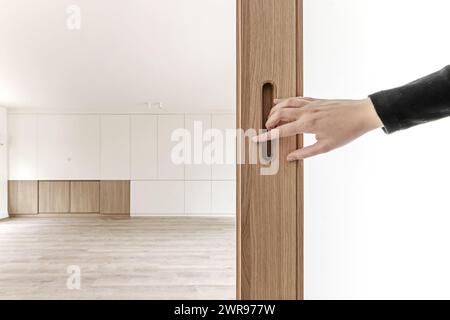 Une main ouvrant une porte sur un salon vide d'une maison individuelle à plusieurs étages avec des planchers en bois de qualité et un mur avec quatre fenêtres en aluminium blanc Banque D'Images