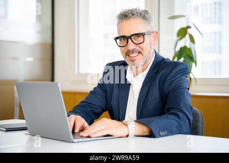 Amical et positif employé masculin mature ou gestionnaire utilisant un ordinateur portable, homme d'affaires senior dans l'usure formelle et le typage de lunettes, répondant aux courriels assis dans le bureau contemporain Banque D'Images