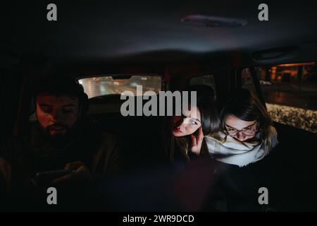 Deux jeunes femmes regardent attentivement l'écran d'un téléphone intelligent à l'intérieur d'une voiture la nuit, avec les visages éclairés par la lueur de l'écran contre l'obscurité. Banque D'Images