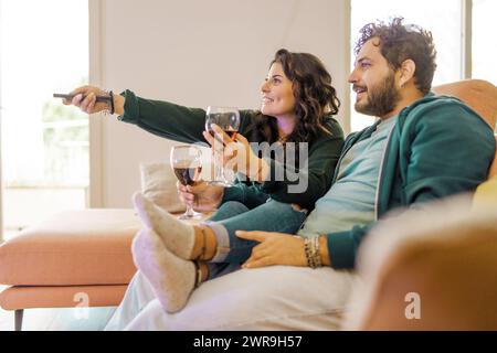 Un couple souriant profite d'une émission de télévision ensemble au cours d'une soirée confortable à la maison, chacun tenant un verre de vin rouge - une image de blis domestique Banque D'Images