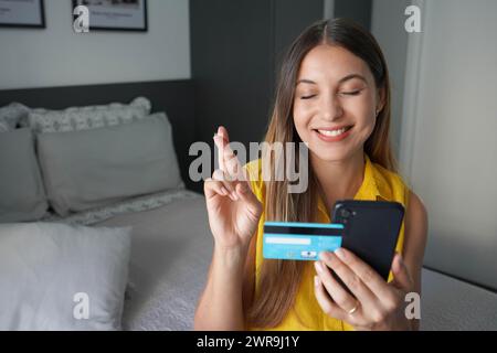 Espoir jeune femme jolie garde les doigts croisés exprime un souhait tient smartphone et carte de crédit attend que les rêves deviennent réalité Banque D'Images