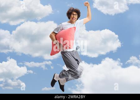 Homme heureux en pyjama tenant un oreiller et sautant haut dans les nuages Banque D'Images