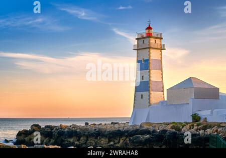 Beau coucher de soleil sur la rive de la mer et phare de Santa Marta à Cascais, Portugal. Ciel coloré, nuages, lumière du soleil, soleil bas, eaux calmes de l'océan, repère local, tour de sécurité et de navigation, rivage rocheux. Banque D'Images