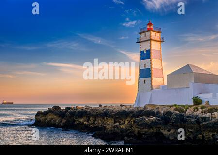 Coucher de soleil coloré sur le rivage de la mer et phare de Santa Marta à Cascais, Portugal. Ciel coloré, nuages, lumière du soleil, soleil bas, eaux calmes de l'océan, repère local, tour de sécurité et de navigation, rivage rocheux. Banque D'Images