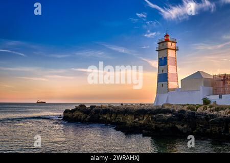 Coucher de soleil spectaculaire sur la rive de la mer et phare de Santa Marta à Cascais, Portugal. Ciel coloré, nuages, lumière du soleil, soleil bas, eaux calmes de l'océan, repère local, tour de sécurité et de navigation, rivage rocheux. Banque D'Images