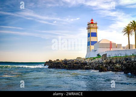 Belle soirée, sur les vagues de marée, les rivages rocheux et le phare de Santa Marta à Cascais, Portugal. Ciel bleu, nuages, lumière du soleil, soleil bas, eaux calmes de l'océan, repère local, tour de sécurité et de navigation, palmiers. Banque D'Images