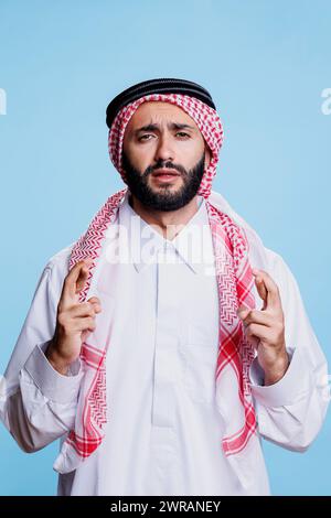 Homme musulman vêtu de vêtements traditionnels faisant le vœu avec les doigts croisés et regardant la caméra avec une expression pleine d'espoir. Arabe portant le thobe priant pour la chance avec un portrait de geste superstitieux Banque D'Images