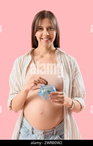 Jeune femme enceinte mettant des pièces dans une tirelire sur fond rose. Concept de prestations maternelles Banque D'Images