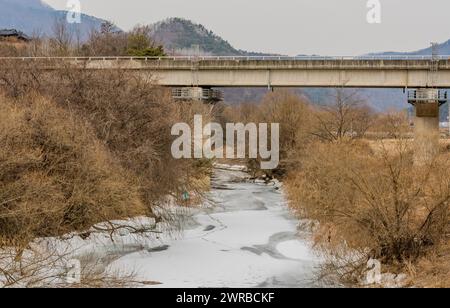 Une scène hivernale couverte montrant un pont sur un ruisseau partiellement gelé avec des arbres sans feuilles, en Corée du Sud Banque D'Images