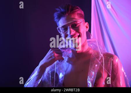 Jeune homme élégant portant un manteau transparent et des lunettes dans les néons Banque D'Images