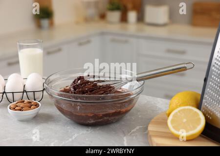Fouet en métal, crème au chocolat dans un bol et différents produits sur une table en marbre gris à l'intérieur Banque D'Images