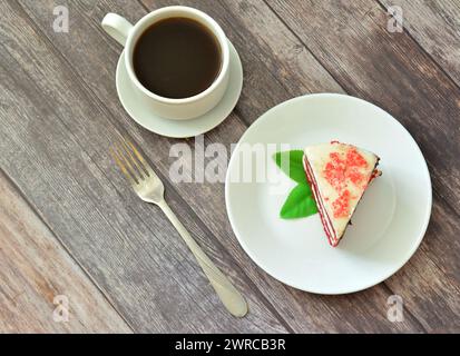 Une tasse de café noir chaud, une fourchette et une assiette avec un morceau de gâteau de velours rouge décoré de feuilles de menthe sur une table en bois clair. Vue de dessus, pose à plat. Banque D'Images