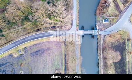 Cette image capture une perspective aérienne d'un canal croisant une route. La photographie montre un passage d'eau contrôlé avec un système d'écluses, permettant aux bateaux de naviguer entre différents niveaux d'eau. La zone environnante est un mélange d'arbres arides et de parcelles d'herbe, indiquant une saison d'automne ou d'hiver tardive. La route courbe élégamment autour du canal, et une petite structure éventuellement liée au mécanisme de verrouillage est visible sur le côté droit de l'image. Aucune personne ni aucun véhicule n'est présent, ce qui souligne le calme de la scène. Vue aérienne d'un canal et d'une intersection de route. Q élevé Banque D'Images